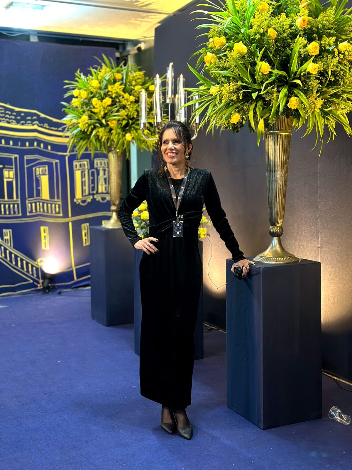 Luciana Jung em evento, vestindo produção preta elegante entre arranjos florais